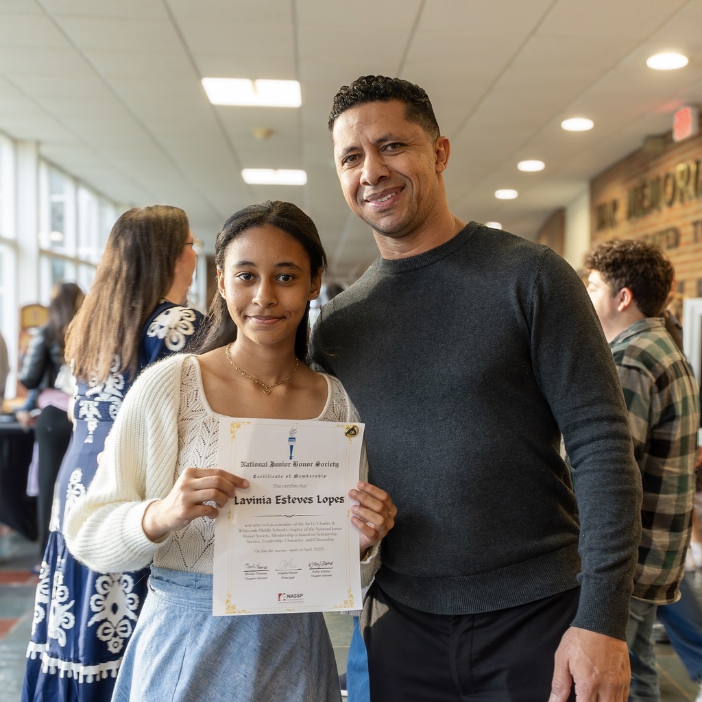 A young female student holds a certificate and stands next to a smiling man in an indoor hallway.
