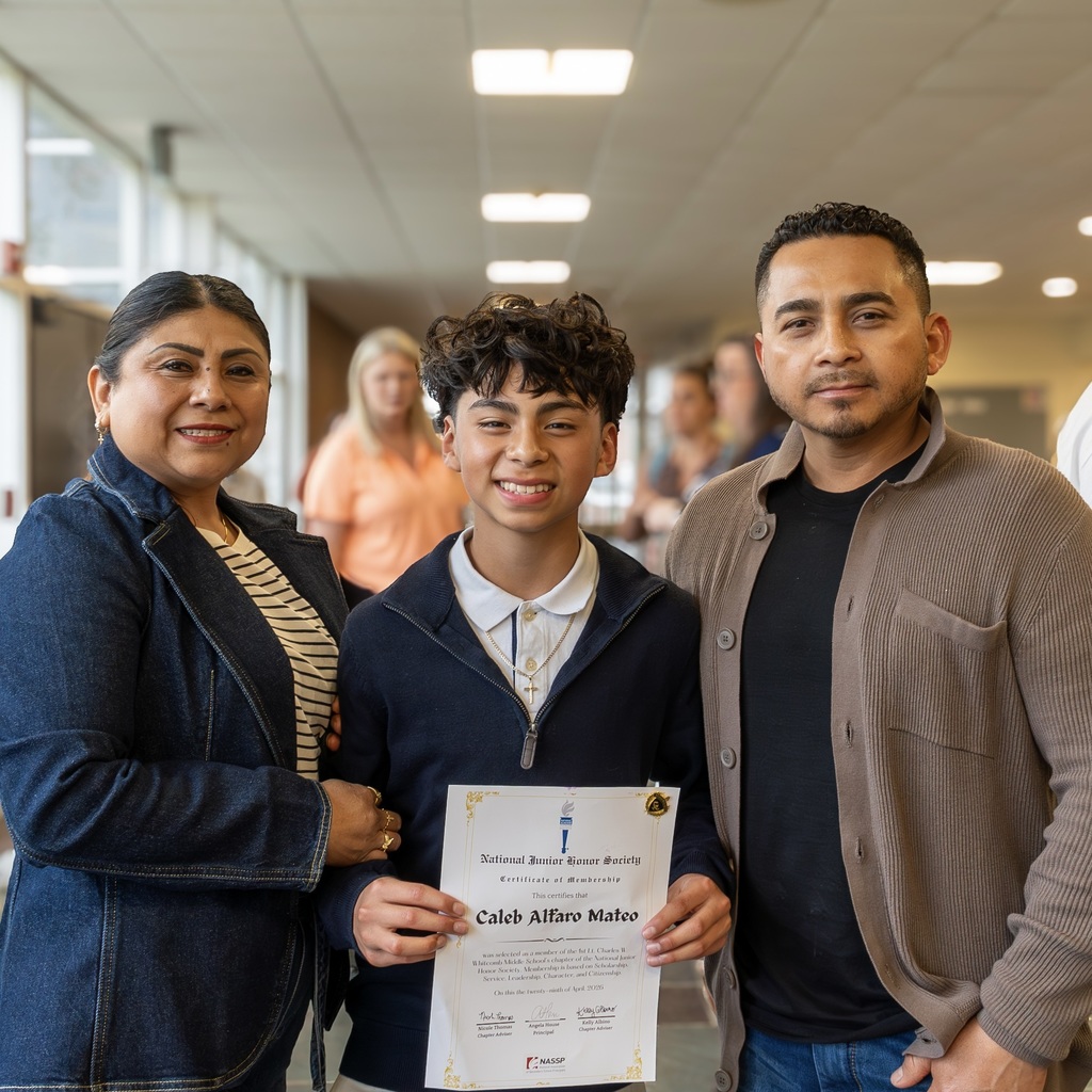 A family stands in a hallway with a student holding a certificate. The woman wears a denim jacket, the male student wears a quarter-zip, and the man wears a brown jacket.