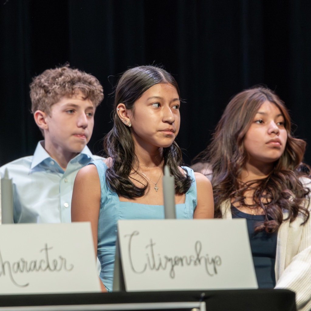 Three students on stage. The words "Character" and "Citizenship" were displayed. One in a blue shirt, another in a white jacket, and the third in a dark top.