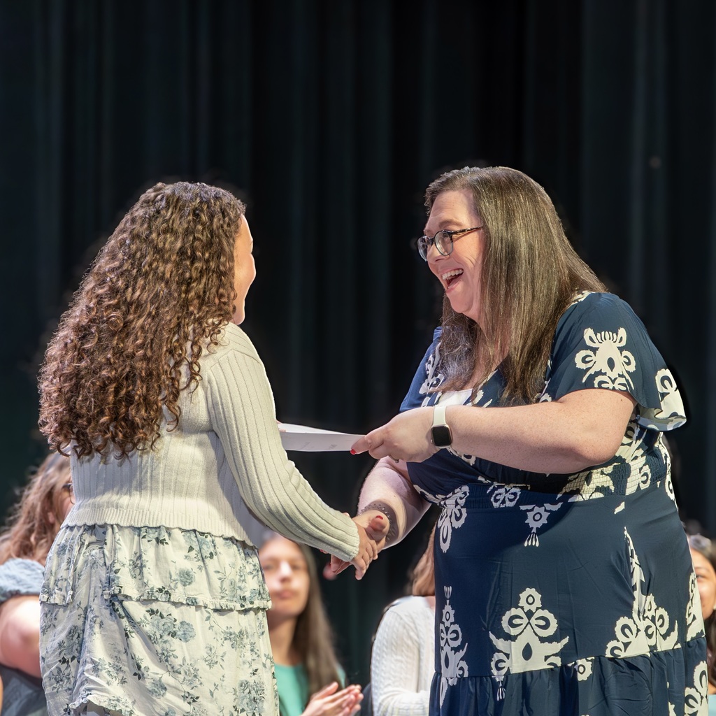 NJHS Adviser Nicole Thomas smiles as she hands a student a certificate during the 2026 NJHS induction. A black curtain and students are seated in the background.