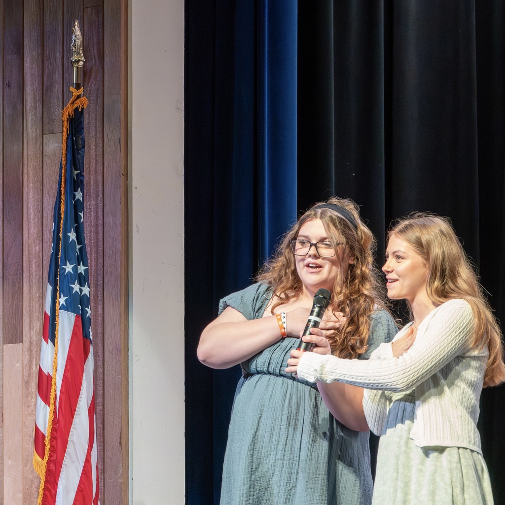 Two Whitcomb students stand on stage, holding onto a microphone. The woman on the left wears glasses and a bracelet.