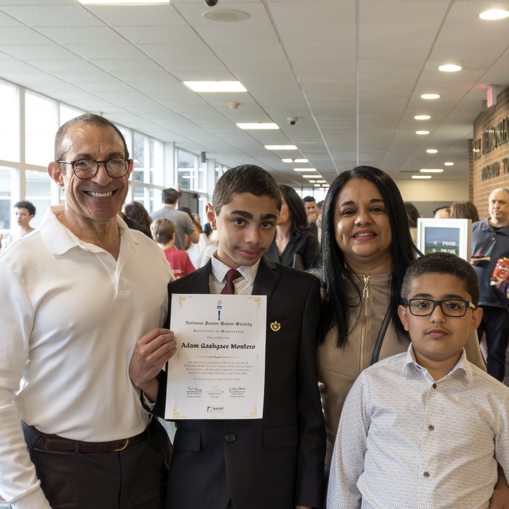 A student holds a certificate in his hand, posing with his family; man in white, a boy in white, woman in light beige.
