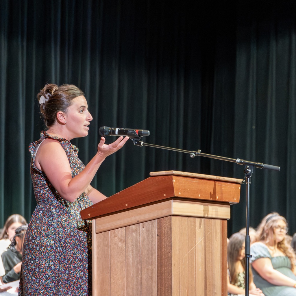 Whitcomb Middle School Principal Angela House is speaking into a microphone at a podium. Behind her, a dark background and students are seated.