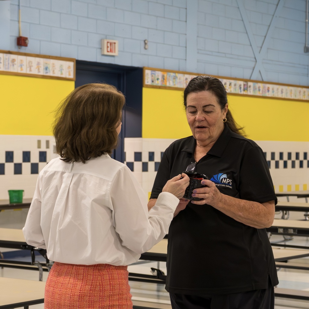 HR Director Patty Brown and a cafeteria worker face each other in a school cafeteria. They both hold onto the same mug. Behind them, yellow and blue walls.