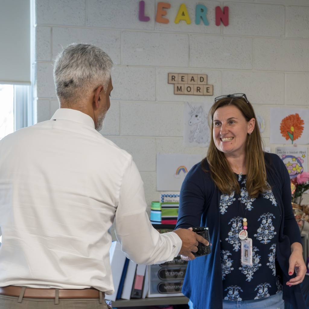 Two people in a classroom: the MPS school business manager, in a white shirt, hands a mug to a smiling staff member in a blue top.