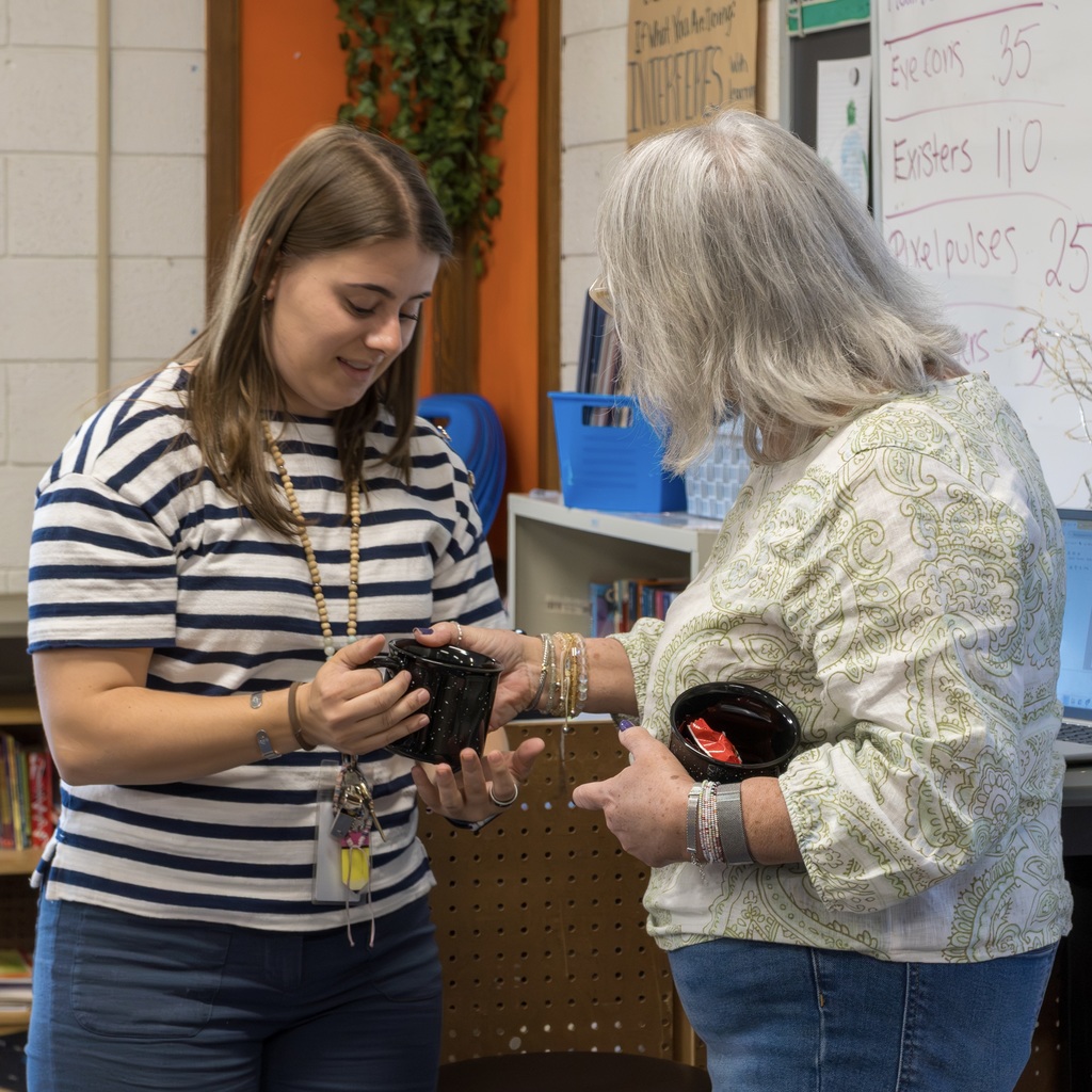 School Committee member Michelle Bodin-Hettinger hands a mug to a smiling staff member, as both stand in a classroom with a whiteboard and books.