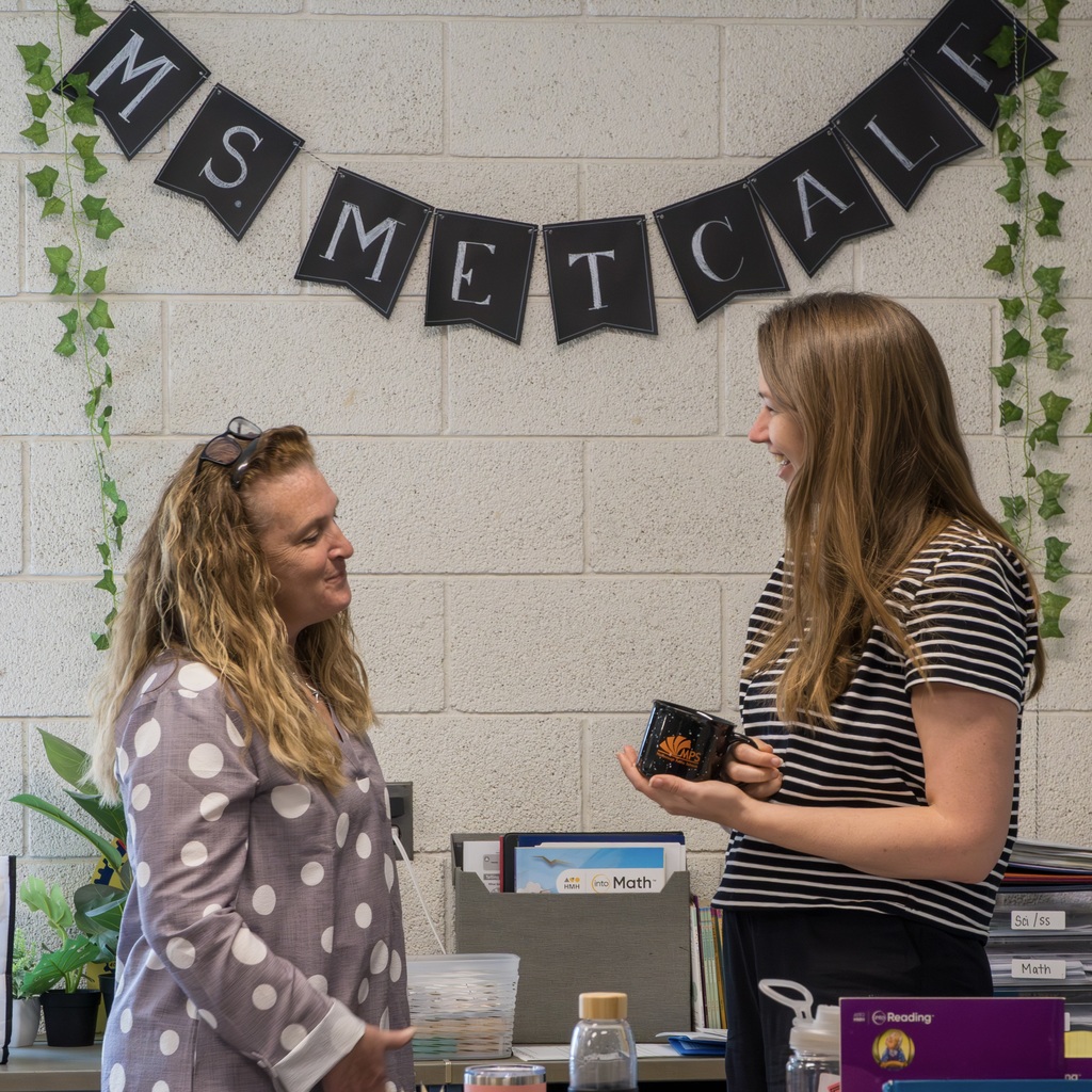 Two women stand facing each other. One holds a small object, the other listens. Behind School Committee members Katherine Hennessy and a Kane School staff member stand facing each other. One holds a black mug, the other listens. Behind them is a banner that reads Ms. Metcalf. is a banner with text.