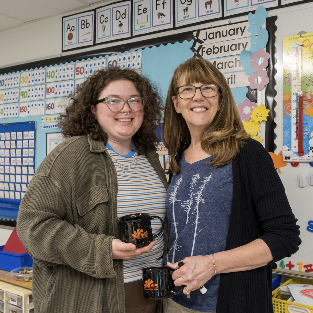 Two Kane School staff members are in a classroom, both holding mugs and smiling. Behind them, a bulletin board shows months and numbers.
