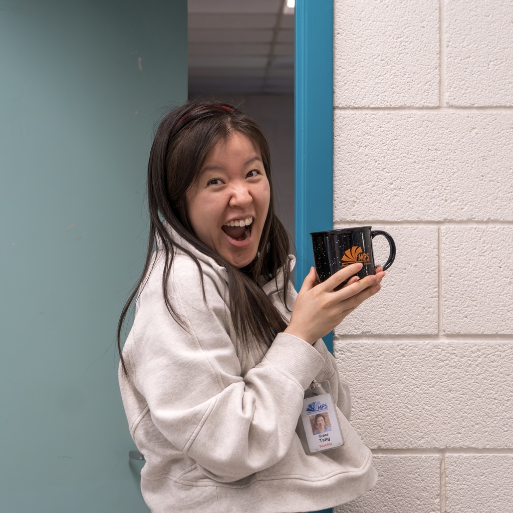A Kane staff member smiling and holding a black coffee mug, wearing a white hoodie and standing against a blue door.