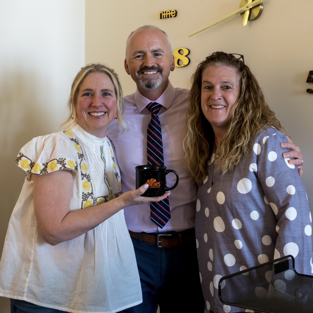 Superintendent Jason DeFalco, School Committee member Katherine Hennessy, and a staff member stand together, smiling. The staff member holds a coffee mug. A clock is on the wall behind them.