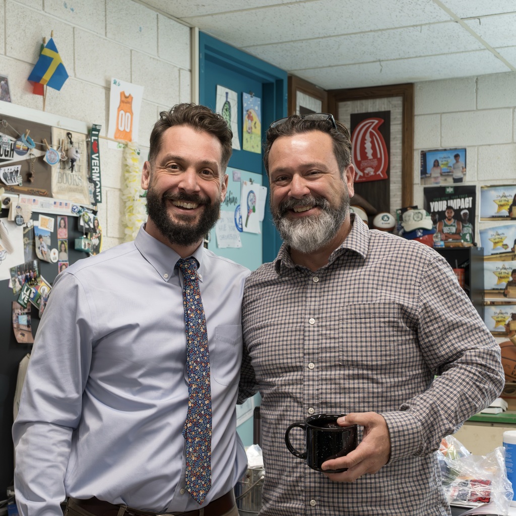 MPS Assistant Superintendent RJ Skaza and a staff member smile and pose. The male Kane School staff member holds a coffee mug. Behind them are bulletin boards with flags and pictures.