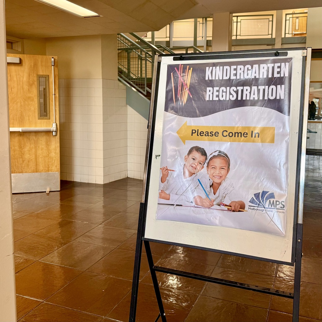 A sign for kindergarten registration stands in an empty room. The sign shows two children. A door and staircase are visible.