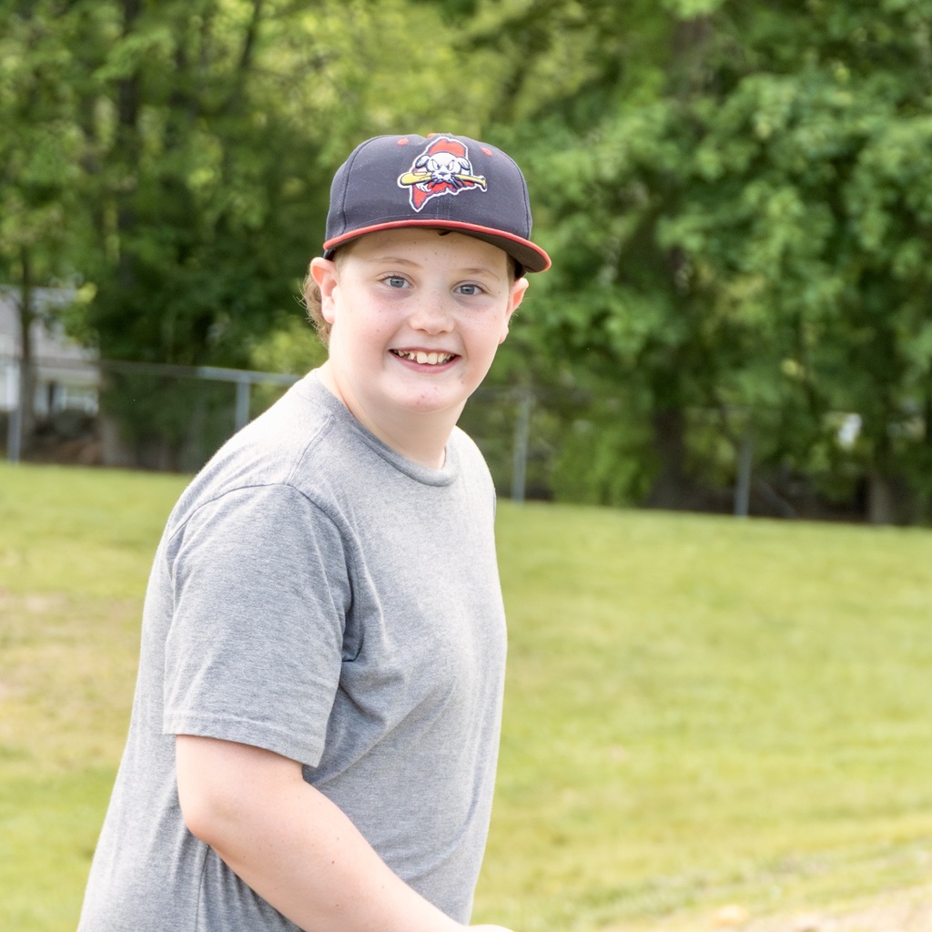 A boy stands in a park, wearing a gray t-shirt and a black hat with white text. Behind him is a grassy area with a metal fence.