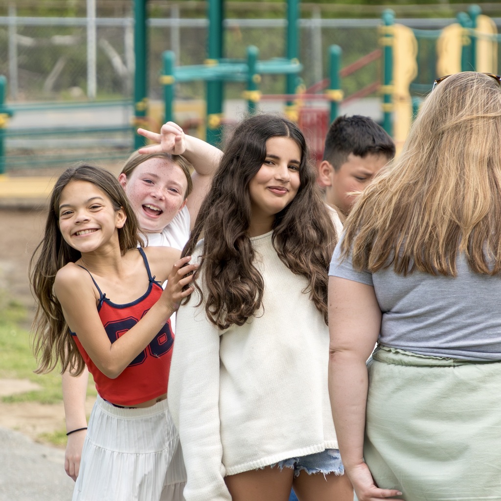 A group of young people stands outdoors near a playground. A girl with long hair smiles, and others look on.