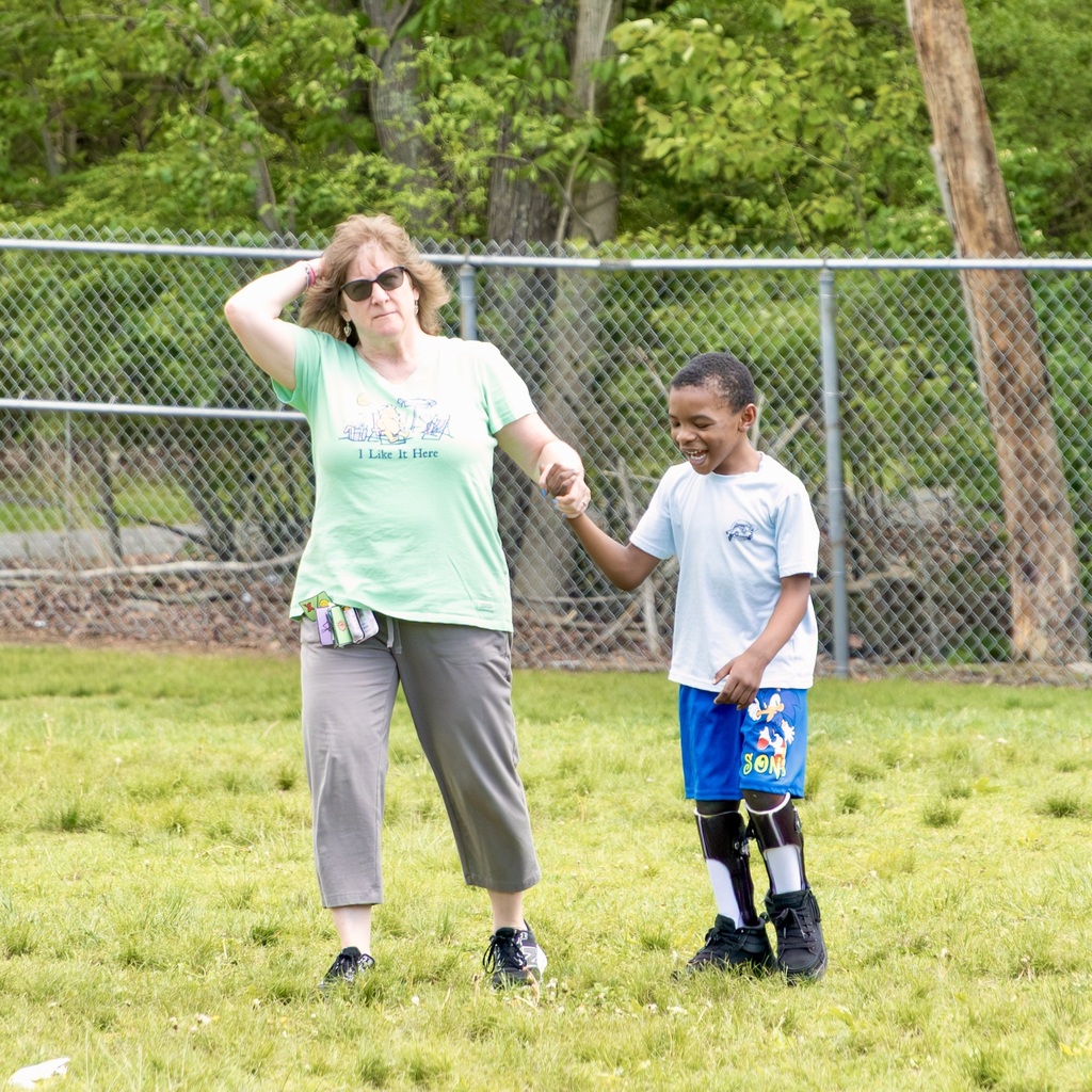 A woman with sunglasses holds a boy's hand in a grassy field with a chain-link fence in the background.