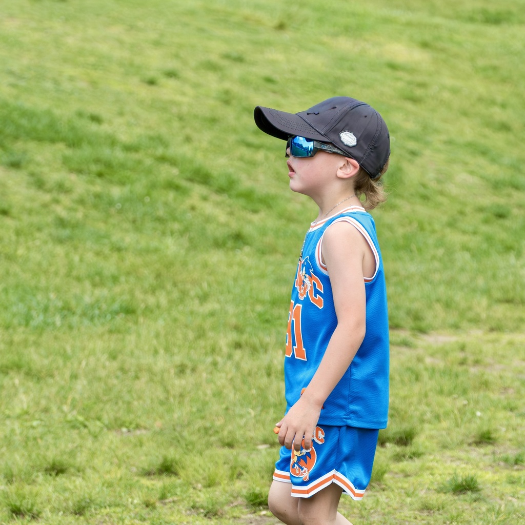 A child in a blue tank top, shorts, and a cap stands on a grassy field, looking upward.