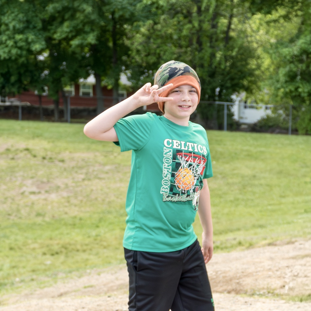 A child in a green t-shirt and a camouflage hat poses for a photo in a park.