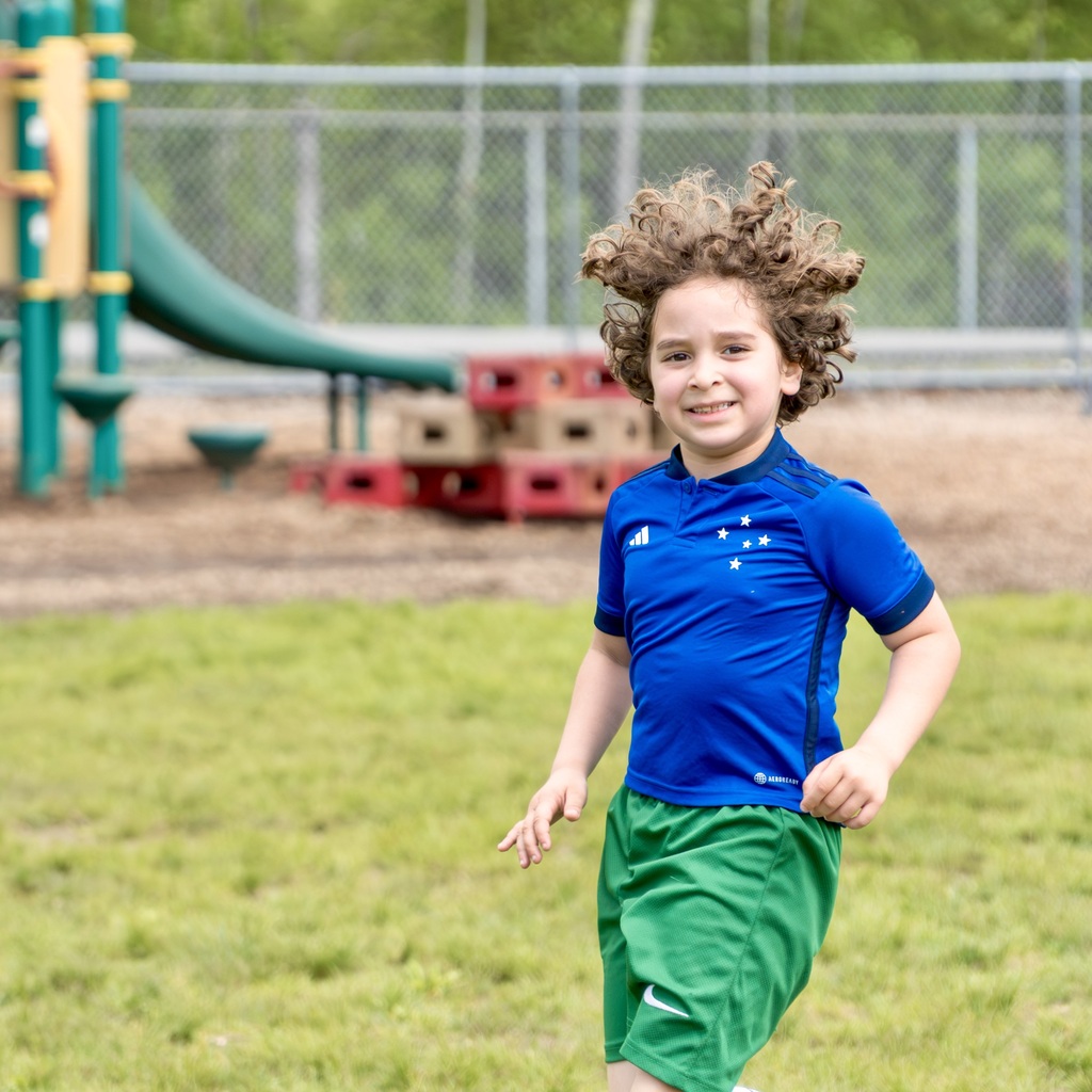 A child with curly hair runs in a park. Behind, a green slide and red blocks are visible.