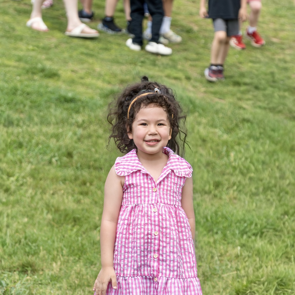 A young girl with curly hair smiles in a pink dress, standing on grass with others behind her.