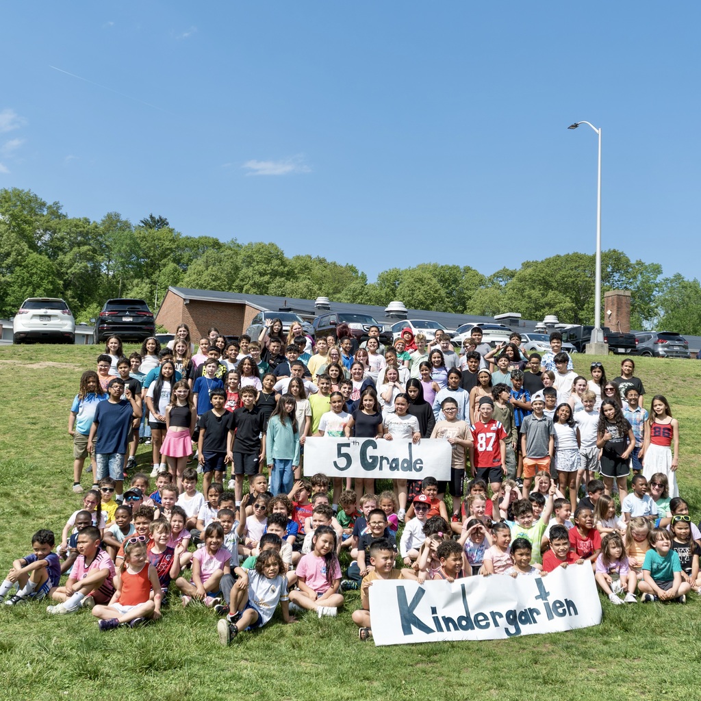 A large group of students in various colors, arranged on a grassy field with two banners reading "5th Grade" and "Kindergarten."