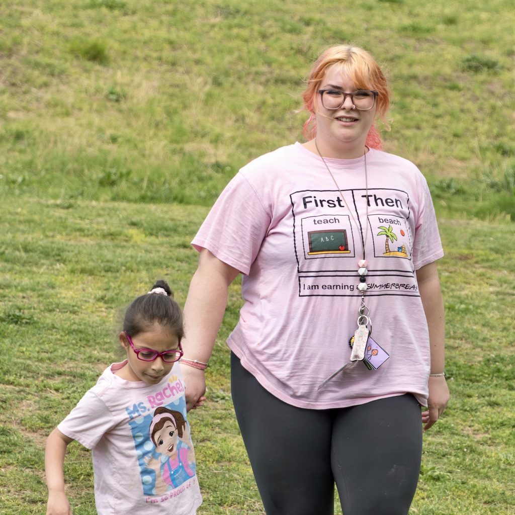 A woman with glasses holds a child's hand; both wear glasses. Woman wears pink shirt, lanyard. They stand on a grassy field.