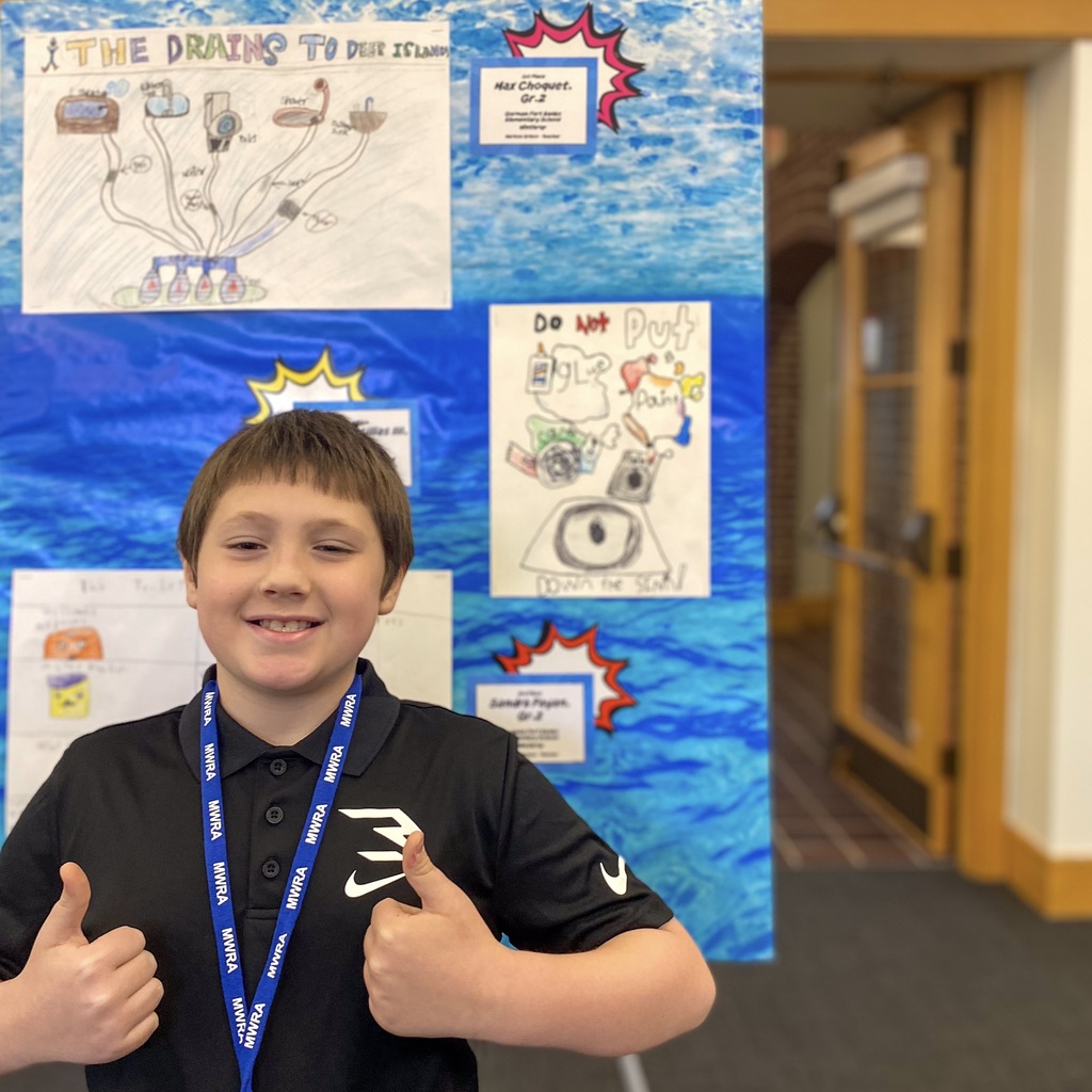 A smiling Richer School boy wearing a blue lanyard and showing two thumbs up signs stands in front of a poster.