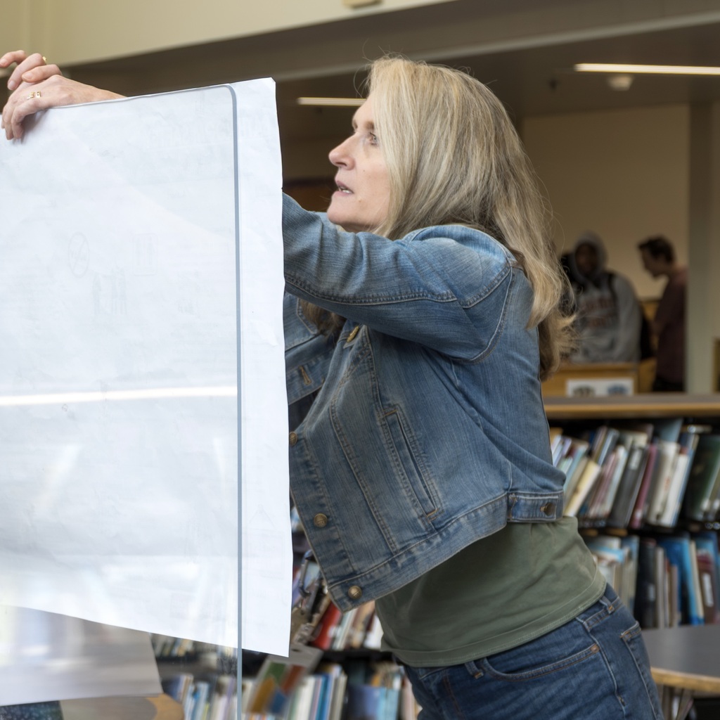 A female Whitcomb staff member holding a large sheet of paper in a room with shelves of books and other people.