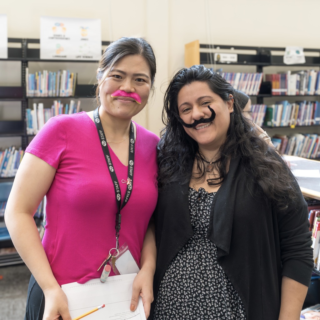 Two Whitcomb staff members with fake mustaches pose together in a library, one holding papers.