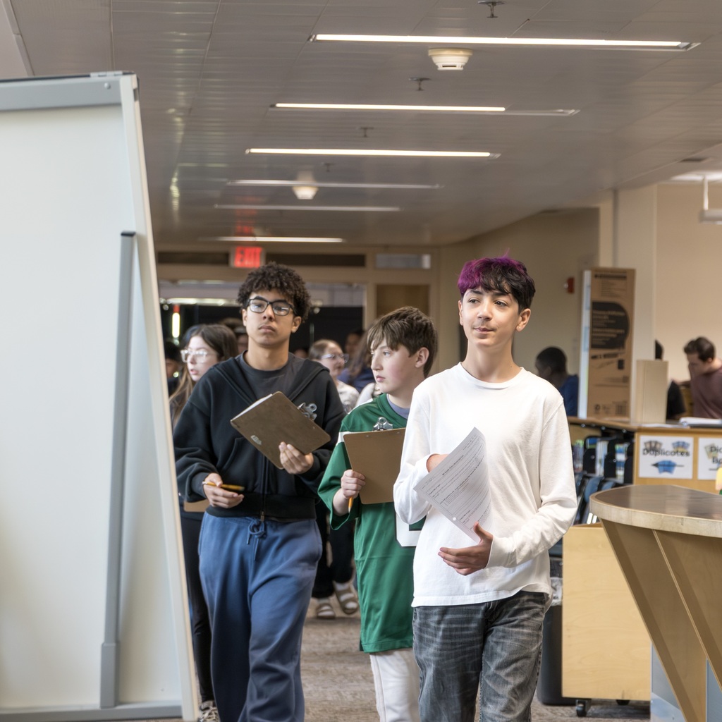 Three Whitcomb students walk down a hallway, two holding clipboards, one holding a paper. They wear casual clothing. Bright overhead lights.