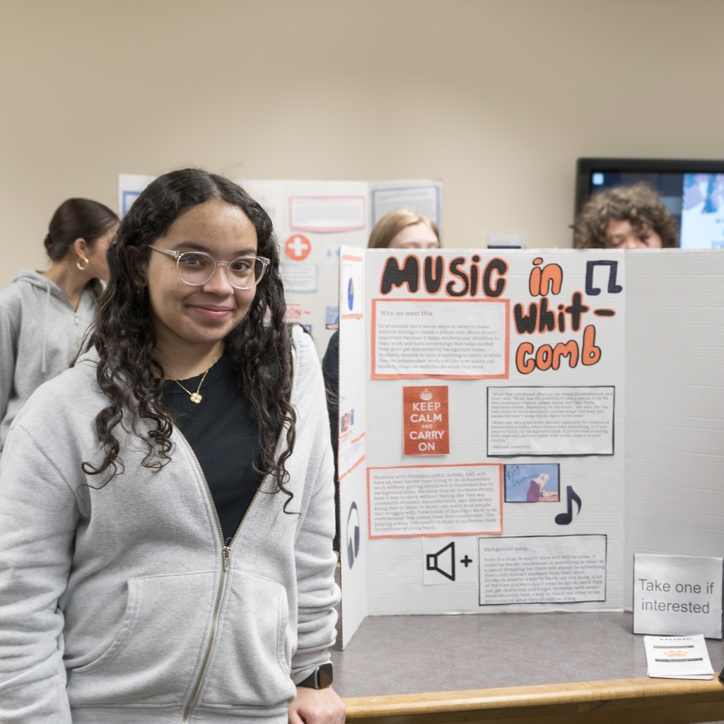 A Whitcomb girl stands before a music poster. Behind her, others view posters. The room is light with tan walls.
