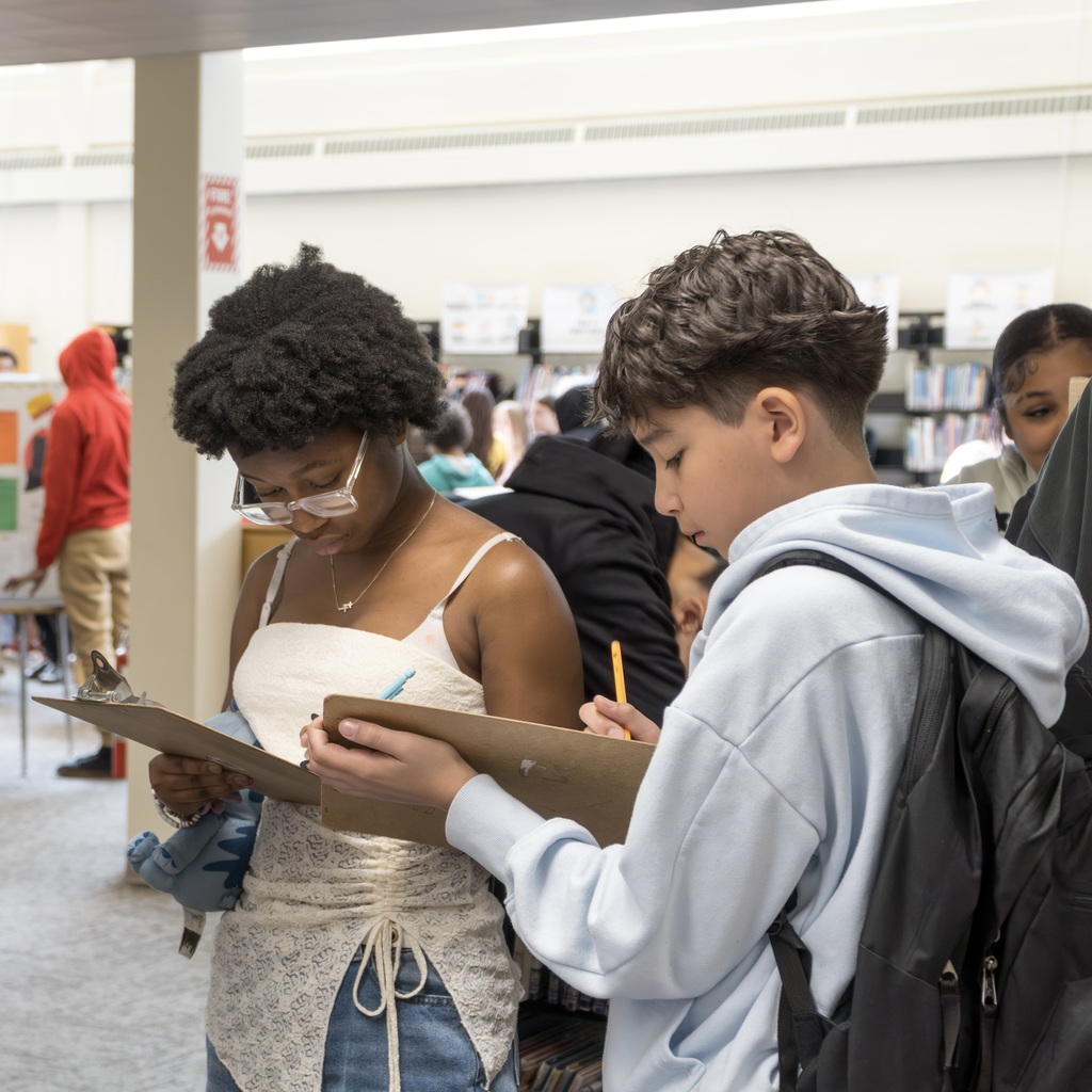 Two Whitcomb students, a girl in a white dressy top and jeans and a boy in a blue hoodie, stand, writing on a clipboard.