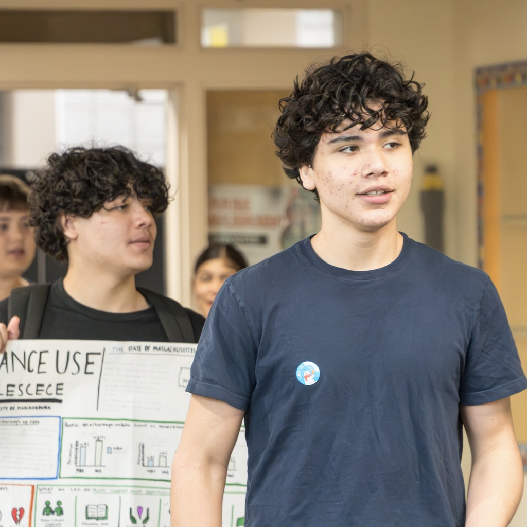 Two Whitcomb students stand in a room. One holds a sign, the other wears a blue shirt. They are both looking forward.