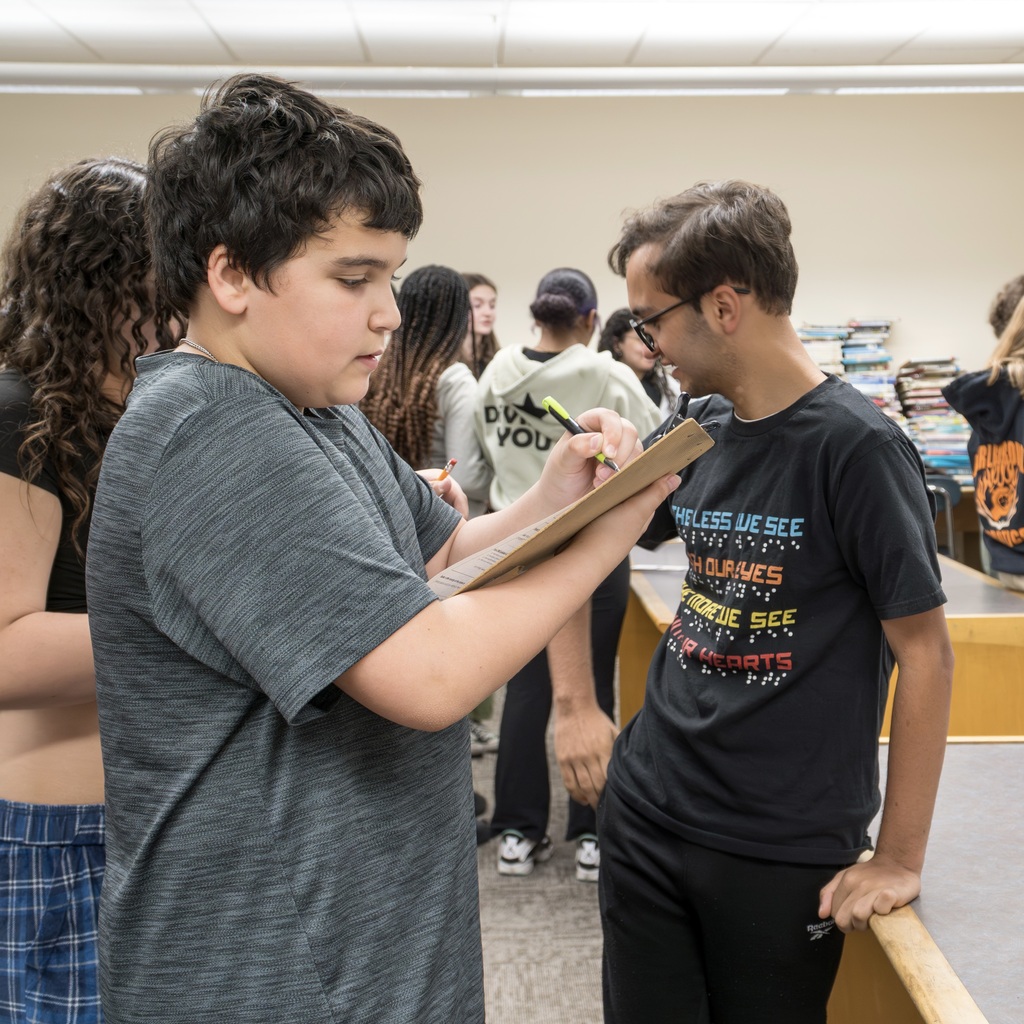 A Whticomb School boy writes on a clipboard while another student looks over his shoulder. Others stand behind them.
