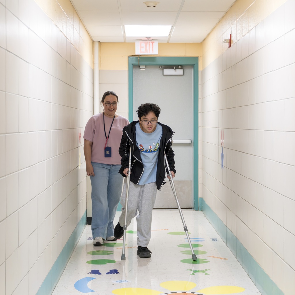 A Jaworek staff member and student walking in a corridor. The student uses crutches. A door and an exit sign are behind them.