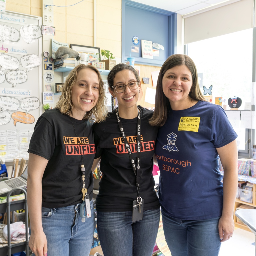 Three women in black and blue shirts stand in a classroom with whiteboards, plants, and a table.