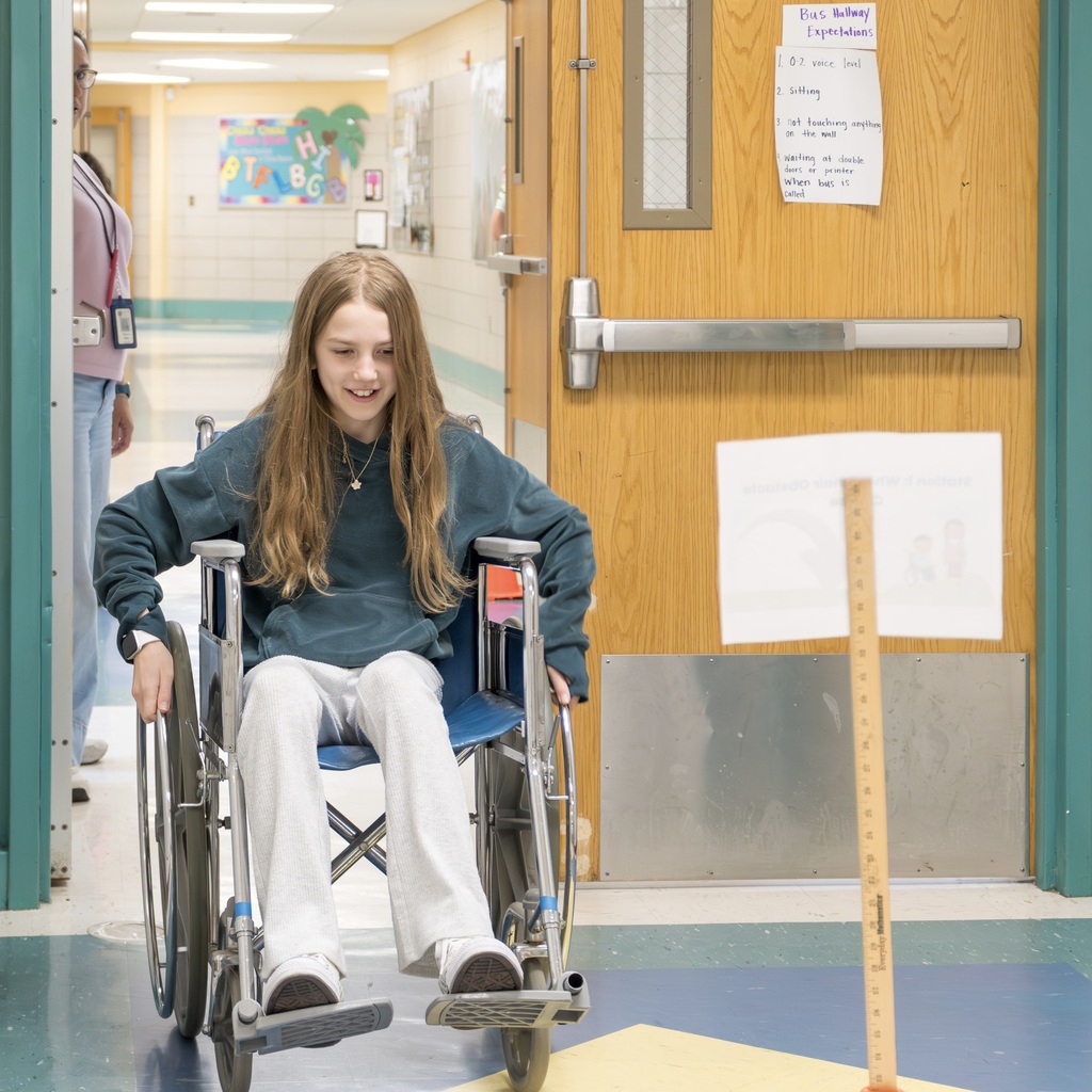 A Jaworek girl in a wheelchair smiles in a hallway, beside a door with a sign and a measuring stick.