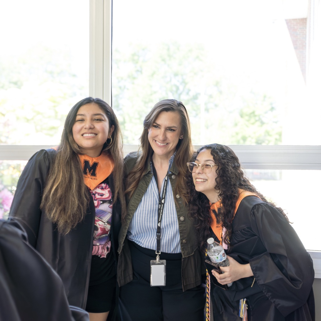 Two Class of 2026 female students in black graduation gowns, and a Richer School teacher, smile in front of a window.