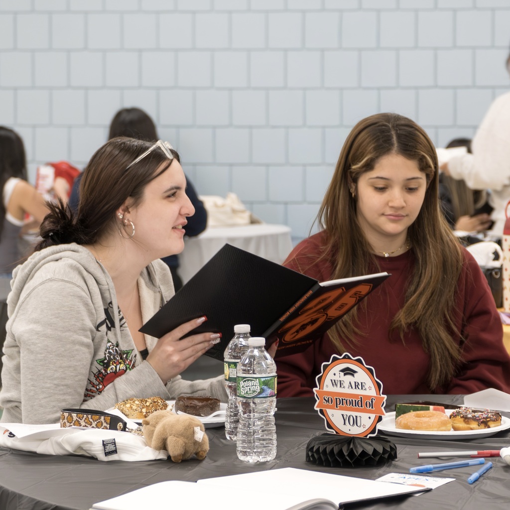 Two Class of 2026 female students sit at a table, reading their yearbooks. They are surrounded by plates, pens, and snacks.