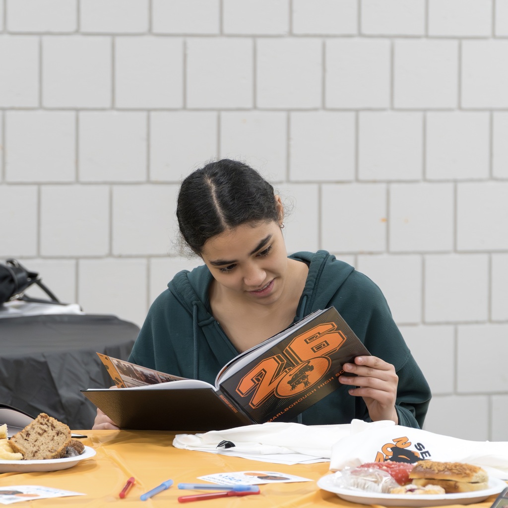 A female Class of 2026 student sits at a table, engrossed in her yearbook. Behind her, a white brick wall. On the table are plates with food, a book, and various items.