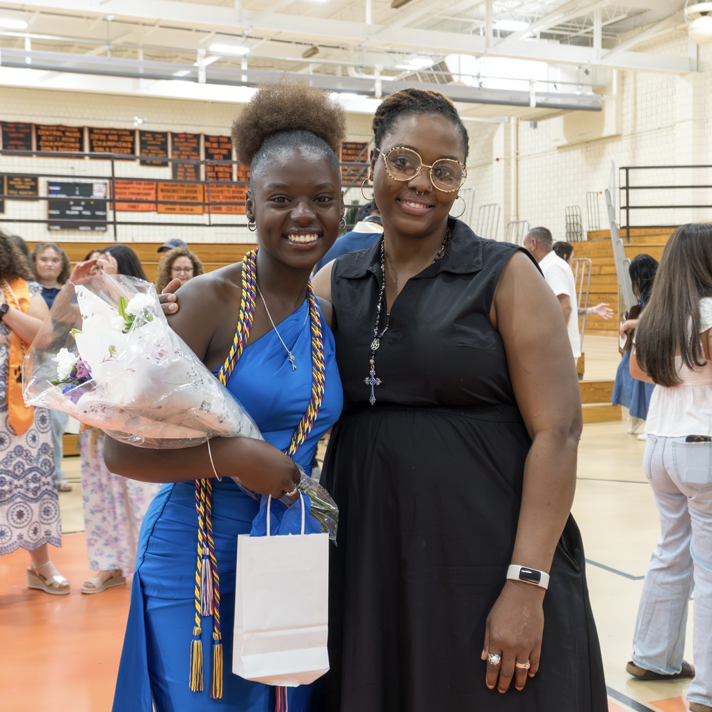 A female Class of 2026 student and a female family member stand next to each other in a gymnasium, with a basketball court visible. One holds flowers and a bag, and the other wears a watch.