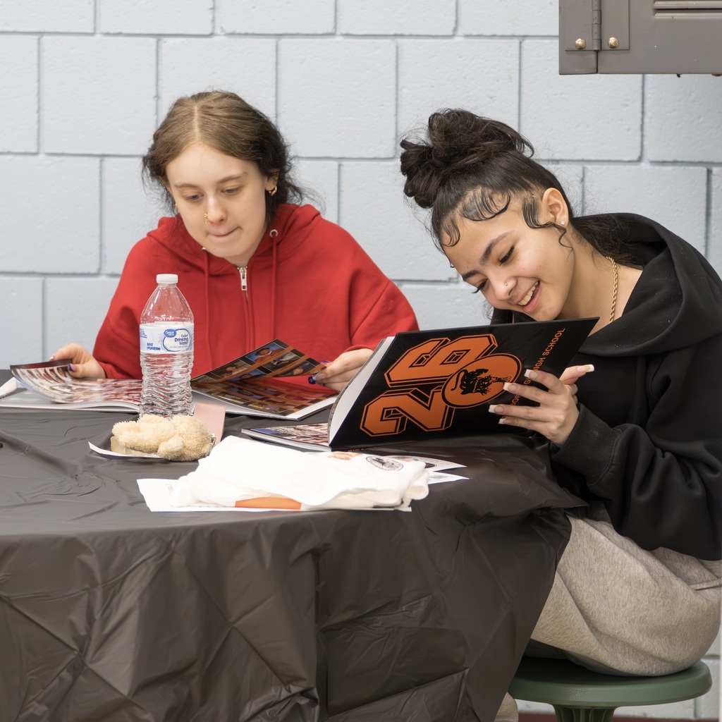 Two MHS students are seated at a table, both reading a yearbook, with food and books.