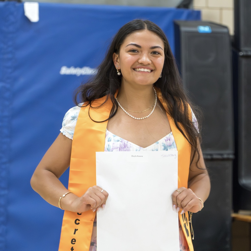 A female MHS student in a graduation cap and gown holding a certificate. She wears a necklace and an orange sash.