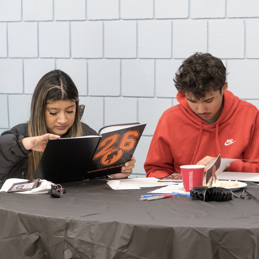 Two MHS students sit at a table, each studying an open yearbook. A female student reads a book with the number "26" on its cover, and the male student studies another book.