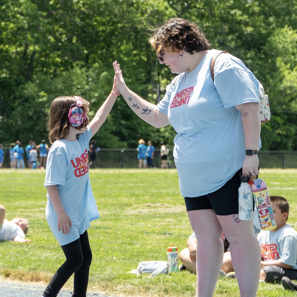 An MPS female staff member and a girl in matching shirts give a high-five. Behind them, a grassy field with people standing and sitting.