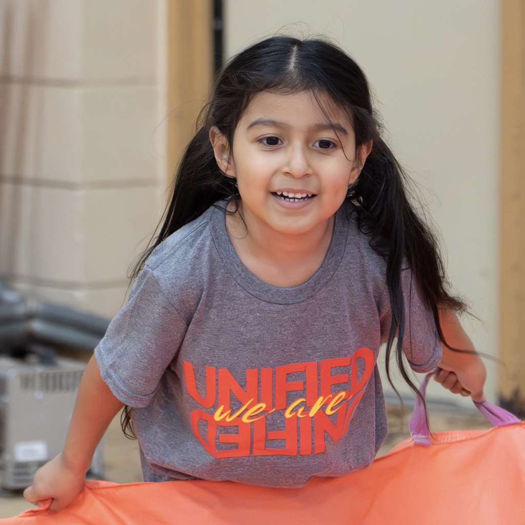 A young girl with long dark hair and a gray shirt with orange letters smiles. She holds part of a large, colorful parachute.