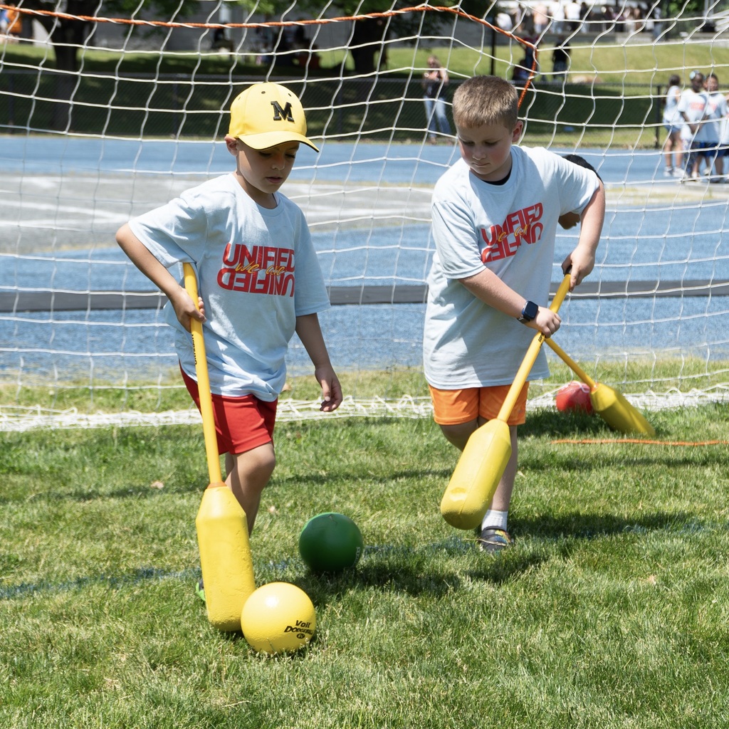 Two children in a field, holding yellow lacrosse sticks, wearing white shirts with red shorts.