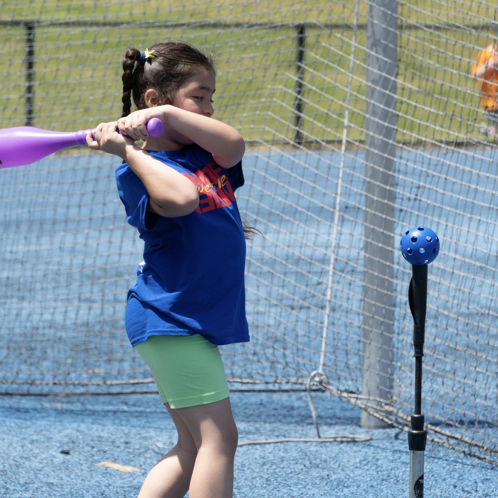 An MPS girl in a blue shirt and green shorts holding a purple bat, standing in front of a net.