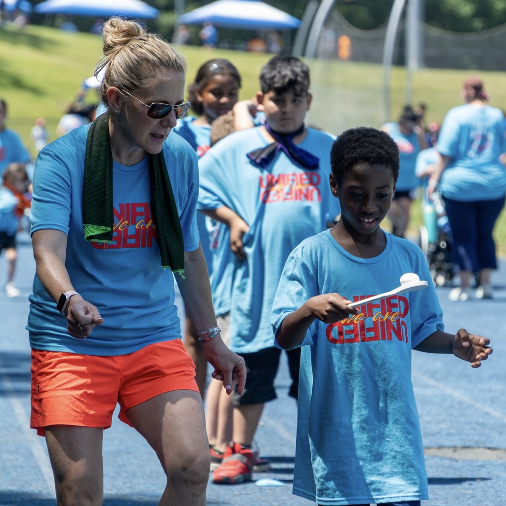 An MPS staff member in orange shorts and a blue shirt walks with a boy attempting the egg drop. They are surrounded by children in blue shirts.