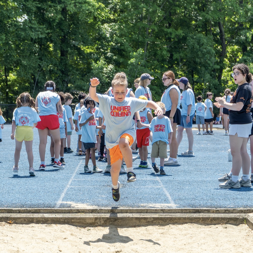 A group of children wearing blue shirts lined up on a track. A student attempts the long jump.