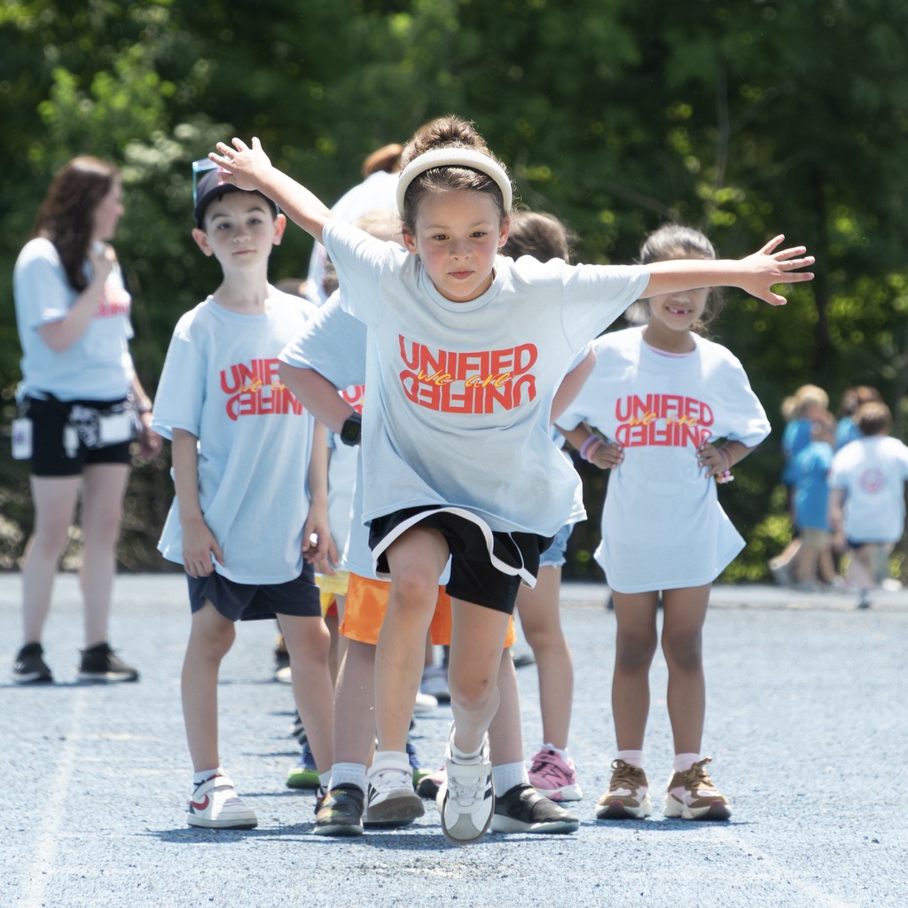 A group of children in matching shirts stands on a track. The child in front jumps up, arms outstretched.